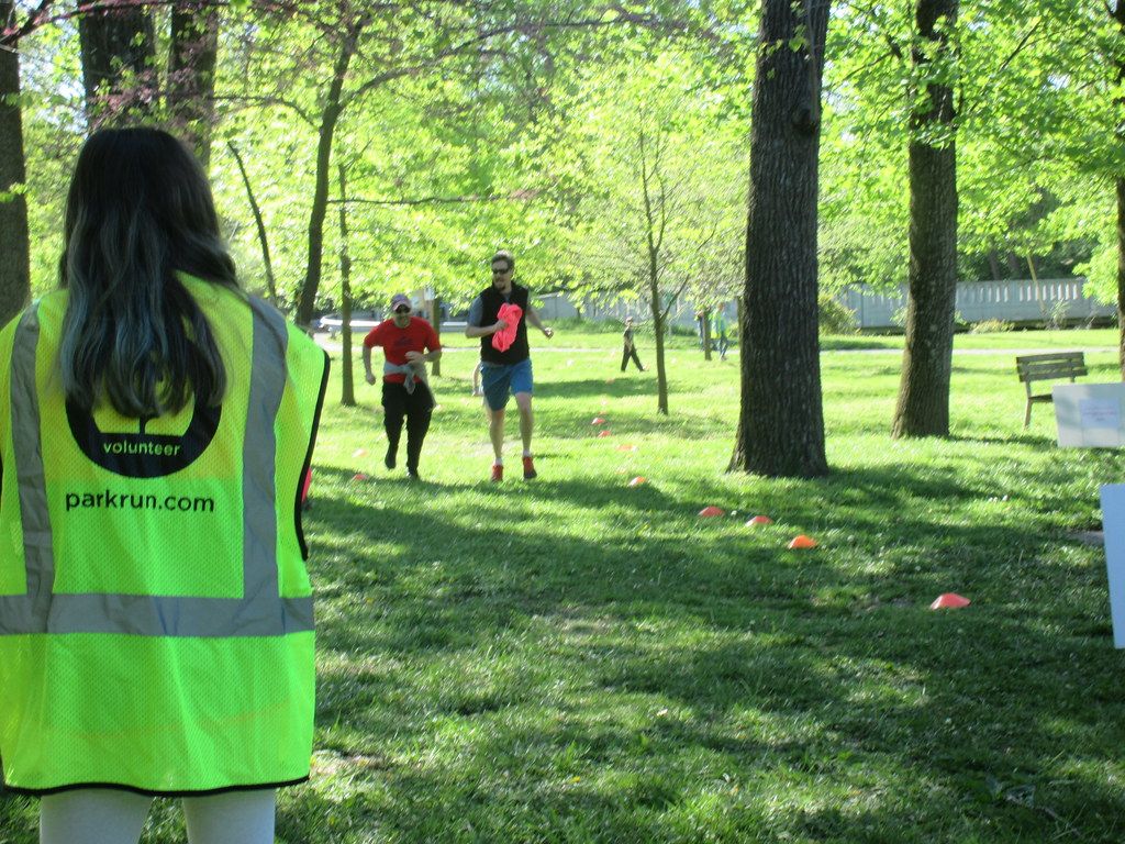 The back of a female parkrun volunteer in hi-vis stoo in a park with two runners running towards her
